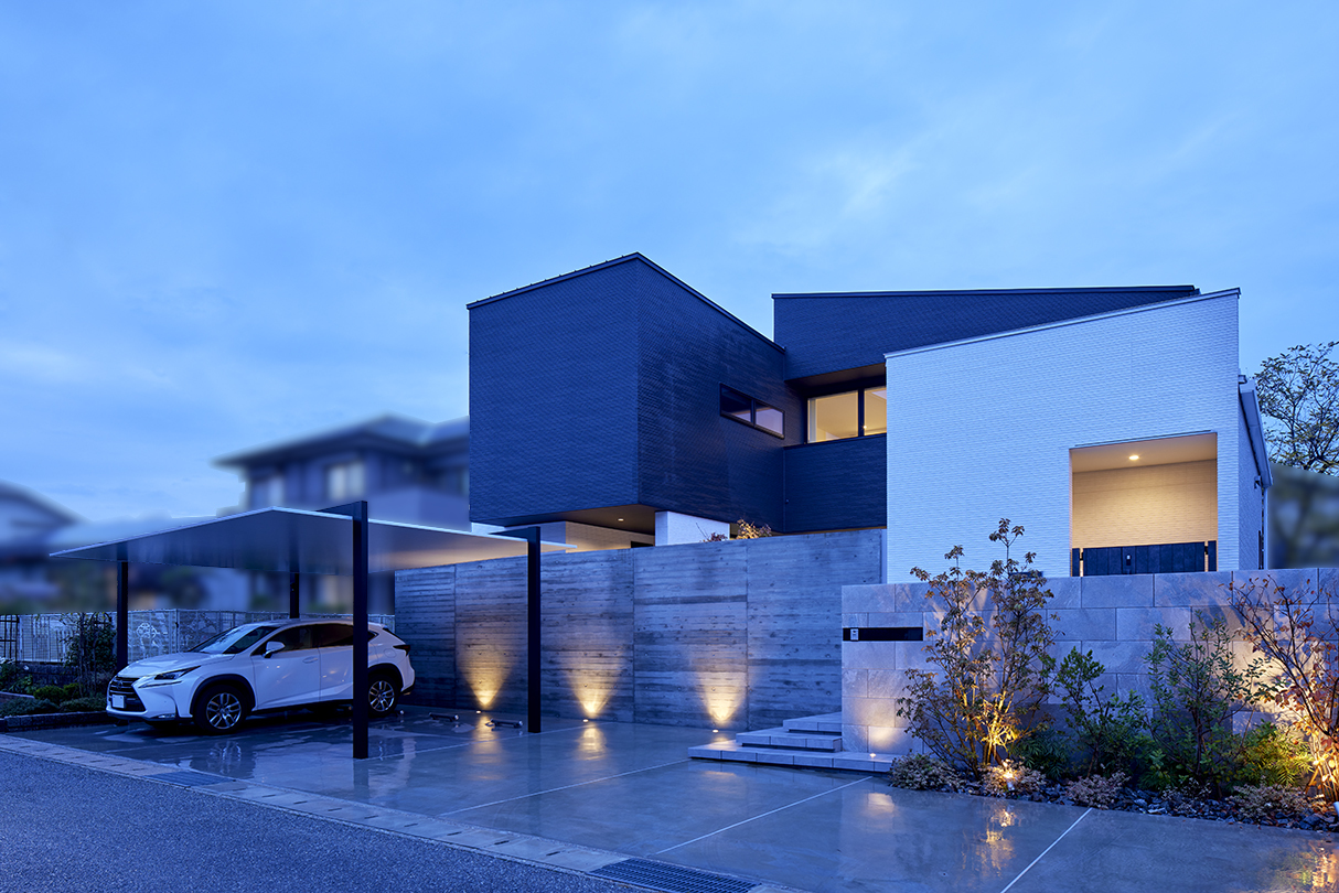夜景のモダンデザイン住宅の外観。黒と白の箱型デザイン、コンクリートの壁とカーポート、間接照明でライトアップされた玄関アプローチ。